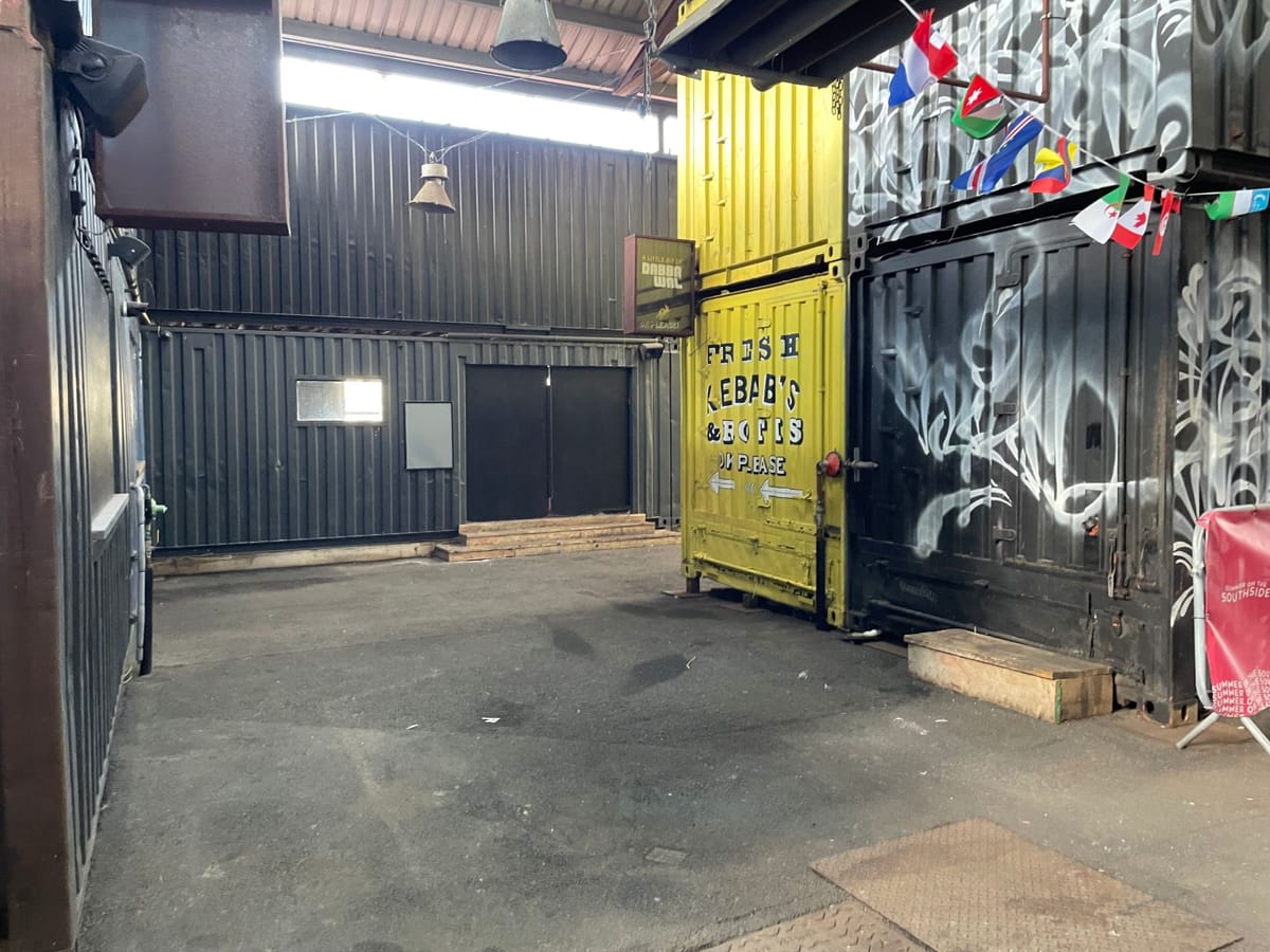 The empty container park in daylight — a covered yard between stacked shipping containers, one painted yellow with a kebab and roti sign, bunting strung overhead.