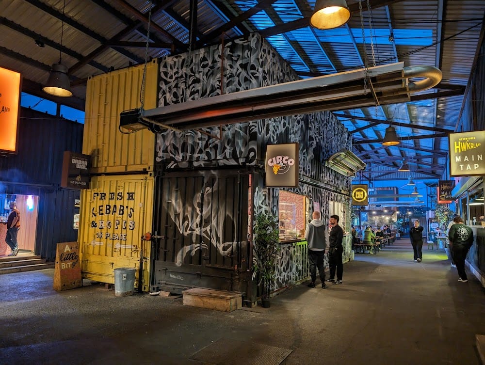 Inside the container park at dusk — stacked shipping containers under a covered walkway, food traders and string lighting.