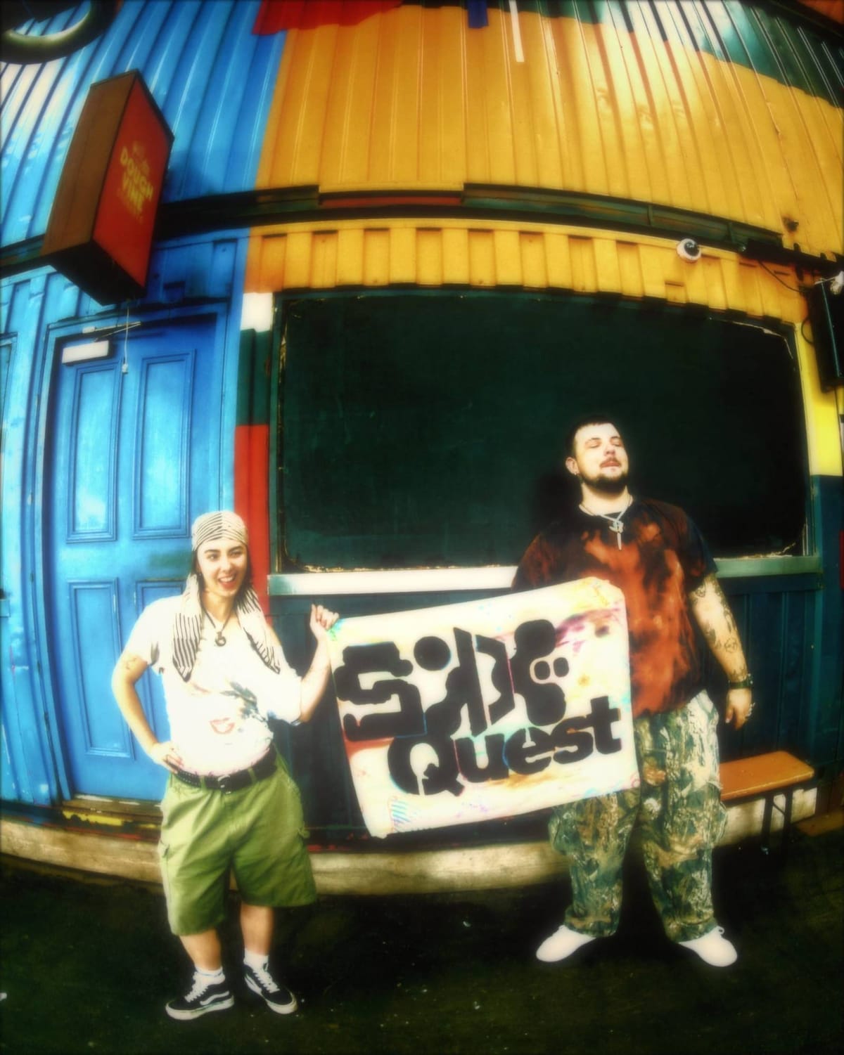 Two of the crew holding a hand-painted Side Quest banner in front of a brightly painted container.