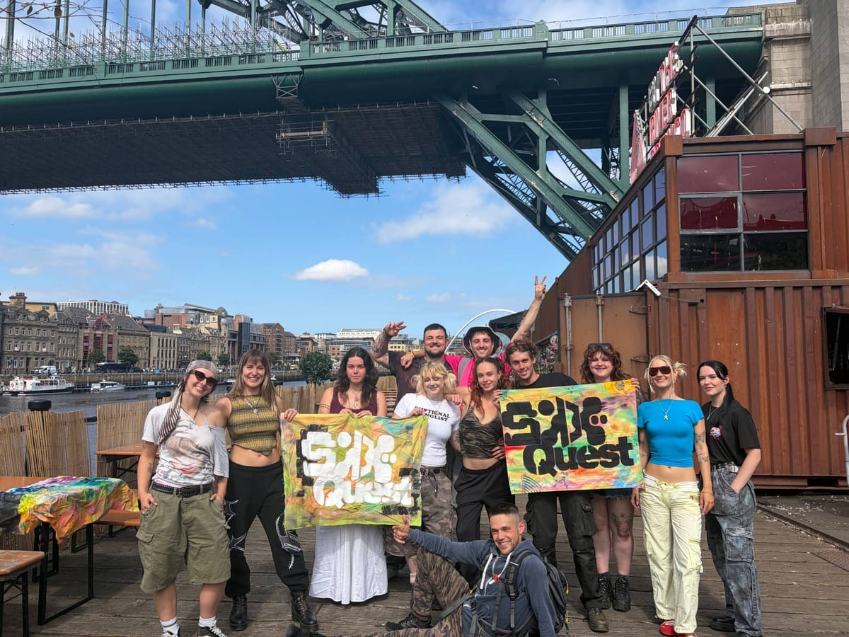 The Side Quest crew on the quayside beneath the Tyne Bridge, holding hand-painted Side Quest banners, with the river and Newcastle quayside behind them.