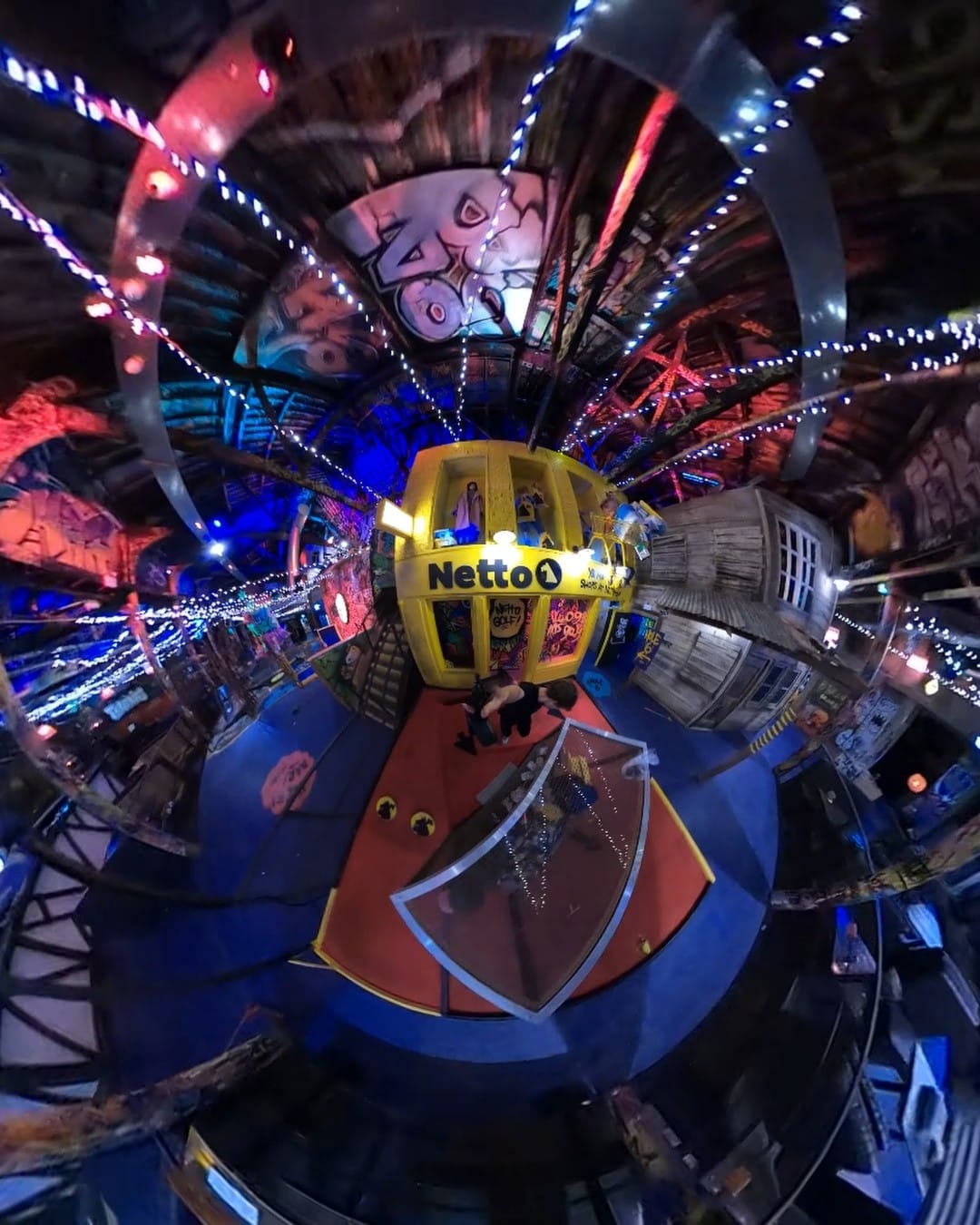 A 360-degree “tiny planet” view of the whole venue, curving the room into a sphere around the yellow Netto Golf kiosk, with strings of lights radiating outward and graffiti-covered walls and rafters wrapped around the edge.